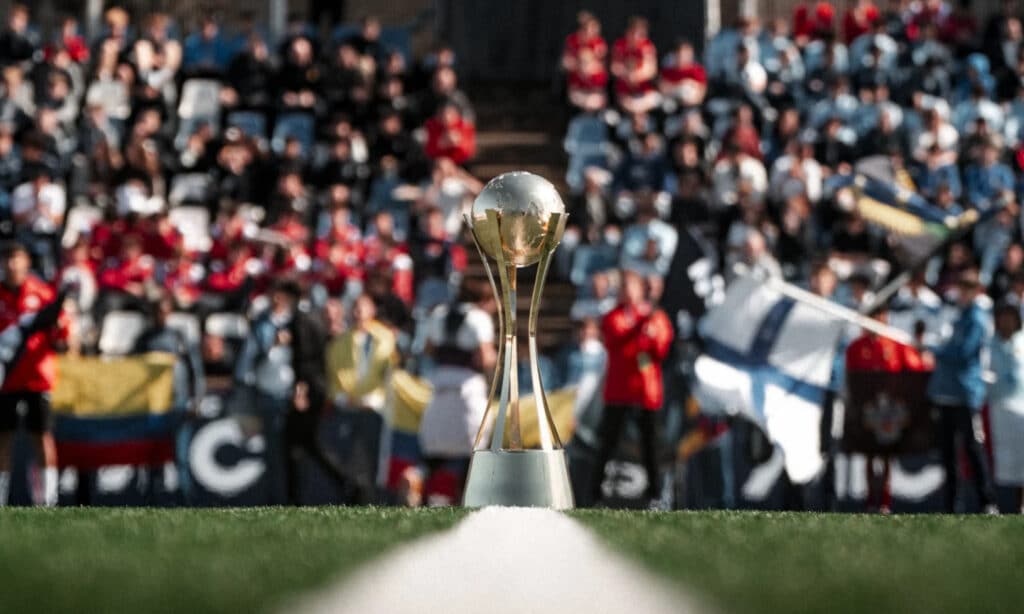 Close-up of the Mediterranean International Cup trophy on the pitch, with fans and flags in the blurred background.