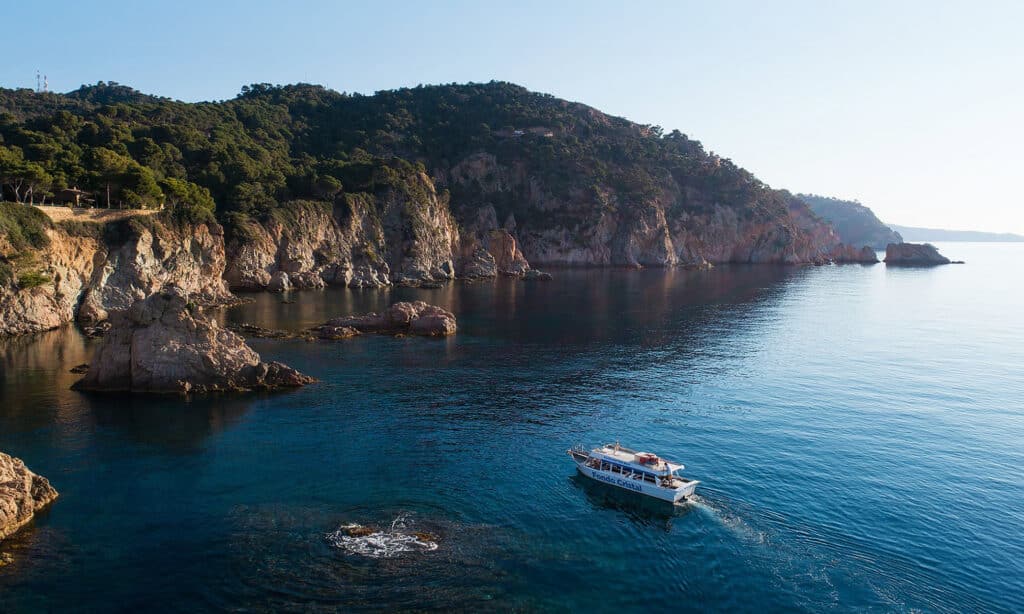 Fondo Cristal glass-bottom boat sailing along the rocky coastline of Tossa de Mar with views of cliffs and crystal-clear Mediterranean waters.
